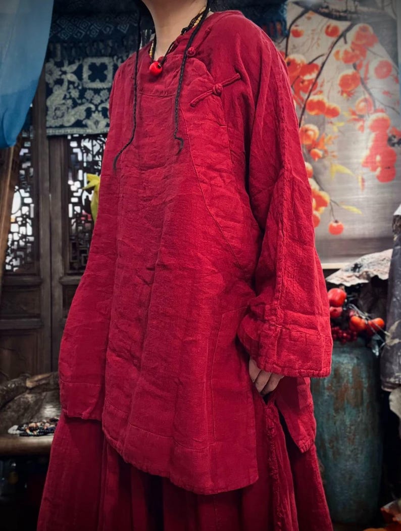 May include: A loose-fitting, long-sleeved, red tunic top with a mandarin collar and a knotted closure at the neckline.