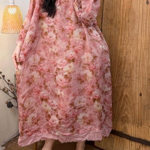 May include: A long, flowing pink dress with a floral pattern. The dress has long sleeves and a round neckline. The fabric appears to be lightweight and slightly sheer. The dress is paired with brown slip-on shoes.