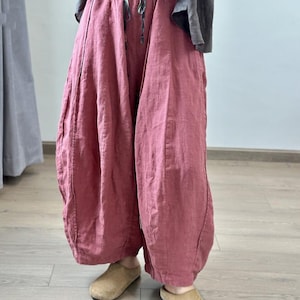 May include: Loose-fitting, dusty rose-colored linen pants with a drawstring waist. The pants have a unique, voluminous shape, gathered at the ankles. The model is wearing tan suede slip-on shoes. The pants are part of a casual, comfortable clothing ensemble.