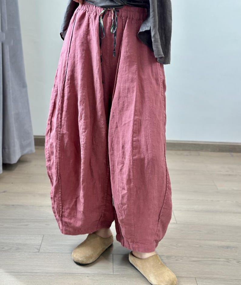 May include: Loose-fitting, dusty rose-colored linen pants with a drawstring waist. The pants have a unique, voluminous shape, gathered at the ankles. The model is wearing tan suede slip-on shoes. The pants are part of a casual, comfortable clothing ensemble.