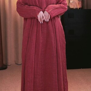 May include: A long, flowing, brick-red dress with long sleeves. The dress features a detailed, embroidered neckline and a loose, comfortable fit. The fabric appears to be a textured, lightweight material, suitable for casual wear.