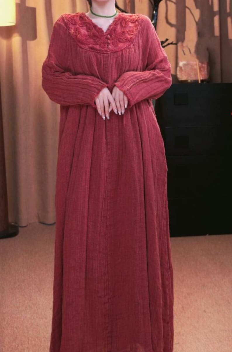 May include: A long, flowing, brick-red dress with long sleeves. The dress features a detailed, embroidered neckline and a loose, comfortable fit. The fabric appears to be a textured, lightweight material, suitable for casual wear.