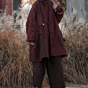 May include: A burgundy quilted coat with a hood and matching pants. The coat has a button closure and patch pockets. The pants are wide-legged. The outfit is worn with tan shoes and is set against a backdrop of tall grasses.