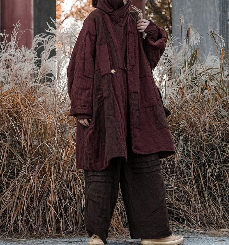 May include: A burgundy quilted coat with a hood and matching pants. The coat has a button closure and patch pockets. The pants are wide-legged. The outfit is worn with tan shoes and is set against a backdrop of tall grasses.