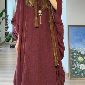 May include: A burgundy-colored, flowing dress with wide sleeves and a matching skirt. The dress features vertical stitching and is accessorized with a long, brown scarf and a silver pendant necklace. The outfit is a bohemian style.