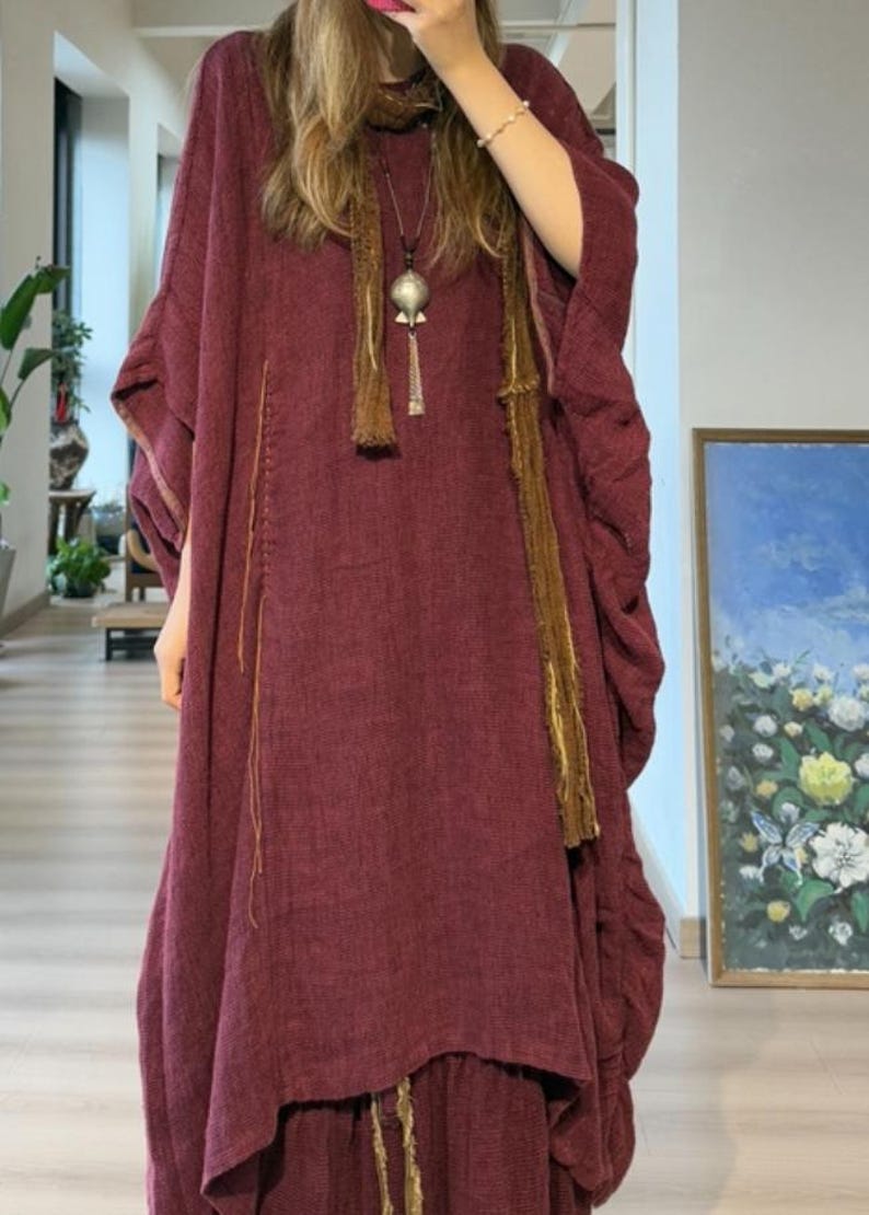 May include: A burgundy-colored, flowing dress with wide sleeves and a matching skirt. The dress features vertical stitching and is accessorized with a long, brown scarf and a silver pendant necklace. The outfit is a bohemian style.