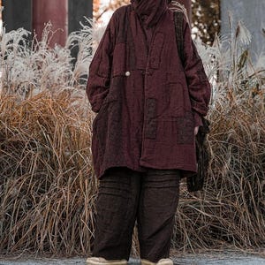 May include: A burgundy-colored, quilted coat with a high collar and a single button closure. The coat has large pockets and is paired with matching dark brown pants. The outfit is worn with tan shoes and a black shoulder bag.