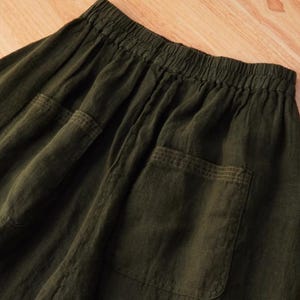 May include: Close-up of olive green linen pants with two large patch pockets. The pants have an elastic waistband and a relaxed fit. The fabric has a slightly wrinkled texture, suggesting a natural fiber.