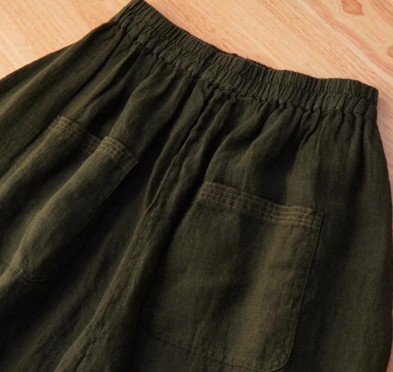 May include: Close-up of olive green linen pants with two large patch pockets. The pants have an elastic waistband and a relaxed fit. The fabric has a slightly wrinkled texture, suggesting a natural fiber.