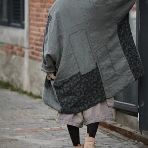 May include: A gray patchwork coat with floral patterned panels. The coat is oversized and appears to be made of linen or a similar fabric. The person is wearing shorts, black tights, and tan boots.