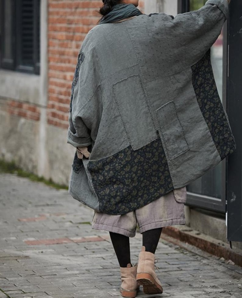 May include: A gray patchwork coat with floral patterned panels. The coat is oversized and appears to be made of linen or a similar fabric. The person is wearing shorts, black tights, and tan boots.