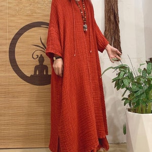 May include: A long, loose-fitting, red,  cotton dress with a V-neck and a drawstring at the neckline. The dress has long sleeves and a flowing skirt.