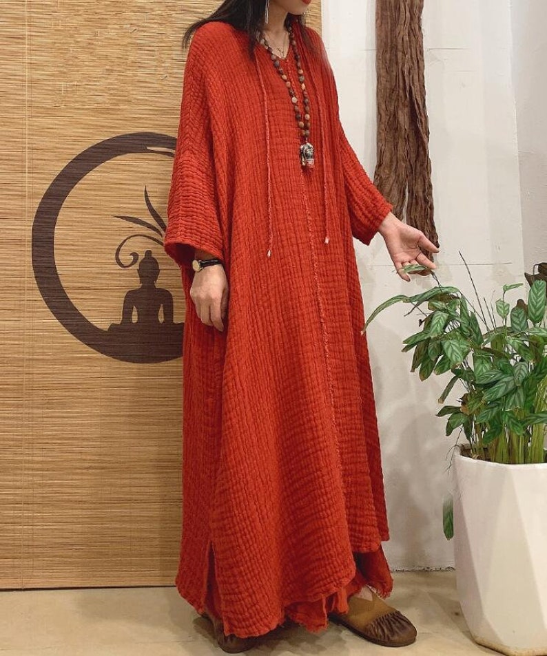 May include: A long, loose-fitting, red,  cotton dress with a V-neck and a drawstring at the neckline. The dress has long sleeves and a flowing skirt.