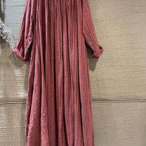May include: A long, flowing dress in a dusty rose color. The dress features a round, embroidered neckline and long sleeves with rolled cuffs. The fabric has a vertical ribbed texture, creating a soft, draped silhouette. A casual, comfortable garment.
