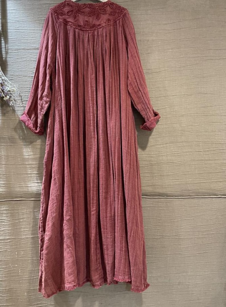 May include: A long, flowing dress in a dusty rose color. The dress features a round, embroidered neckline and long sleeves with rolled cuffs. The fabric has a vertical ribbed texture, creating a soft, draped silhouette. A casual, comfortable garment.