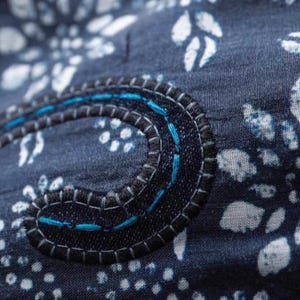 May include: Close-up of a navy blue fabric with a white floral pattern. A dark blue, stitched applique with teal thread accents forms a curved shape. The stitching is detailed and adds texture to the design.