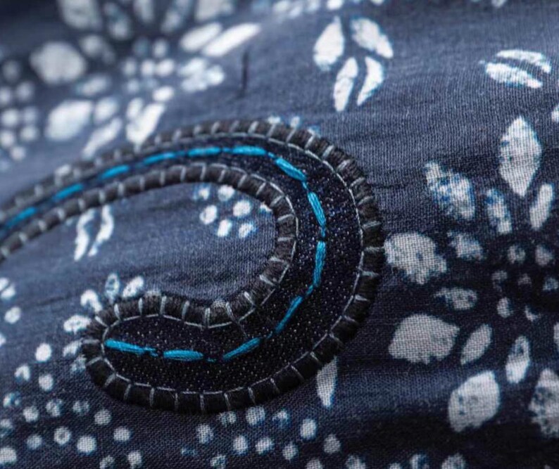 May include: Close-up of a navy blue fabric with a white floral pattern. A dark blue, stitched applique with teal thread accents forms a curved shape. The stitching is detailed and adds texture to the design.