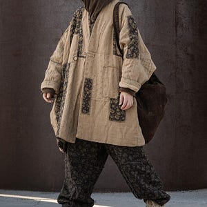 May include: A beige, oversized coat with dark floral patches and matching pants. The coat has a relaxed fit, long sleeves, and a button closure. The pants have a floral pattern. A brown hooded sweatshirt is worn underneath. A large brown bag is carried over the shoulder.