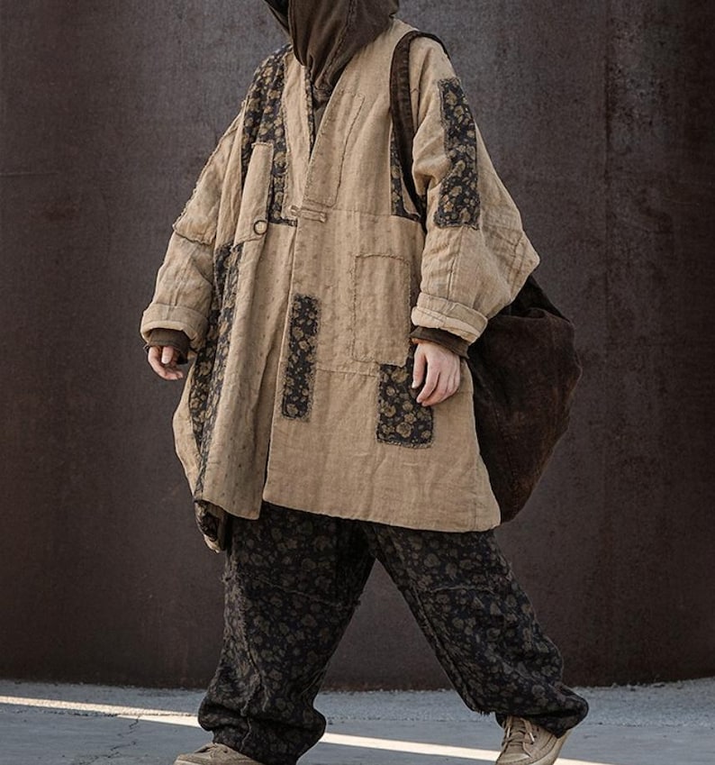 May include: A beige, oversized coat with dark floral patches and matching pants. The coat has a relaxed fit, long sleeves, and a button closure. The pants have a floral pattern. A brown hooded sweatshirt is worn underneath. A large brown bag is carried over the shoulder.