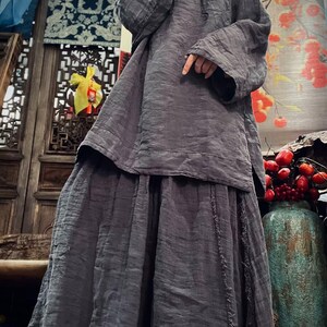 May include: A gray linen tunic top and skirt set with a loose fit and long sleeves. The top has a round neckline and a drawstring closure. The skirt has a tiered design and a raw edge hem.