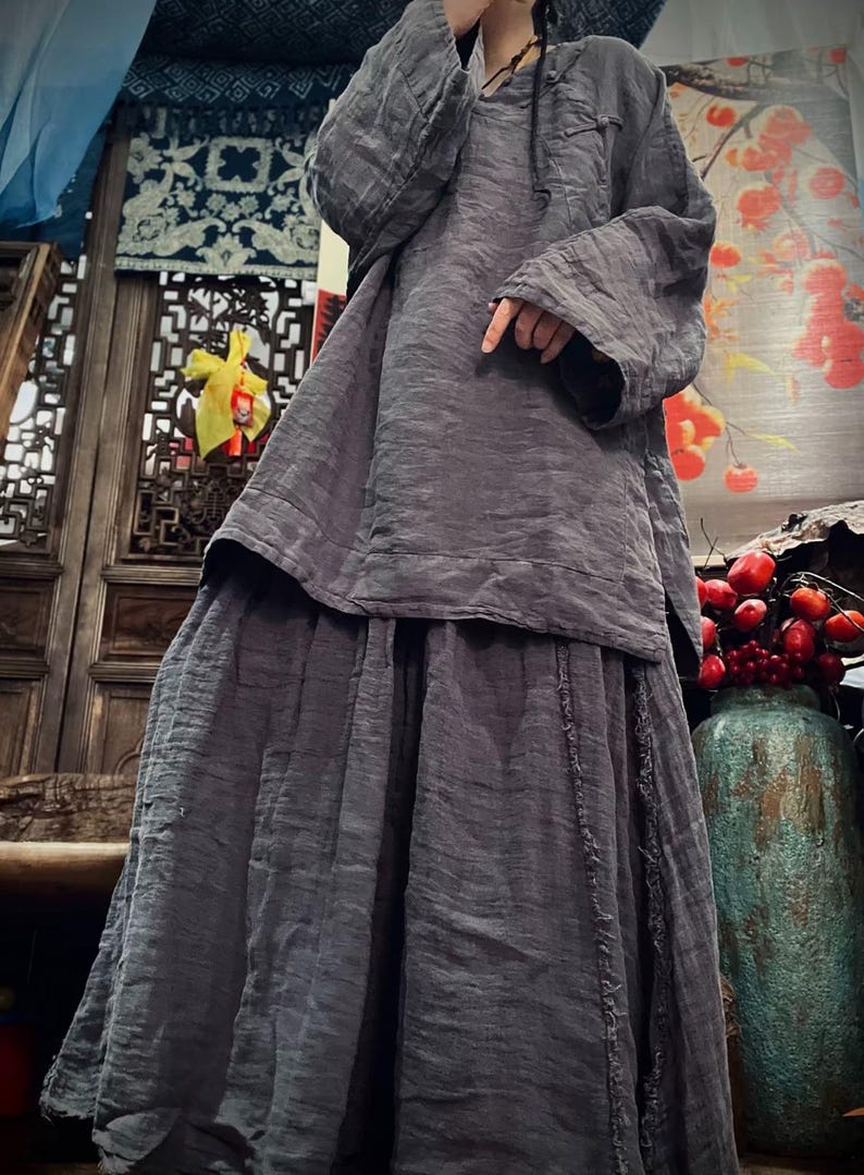 May include: A gray linen tunic top and skirt set with a loose fit and long sleeves. The top has a round neckline and a drawstring closure. The skirt has a tiered design and a raw edge hem.