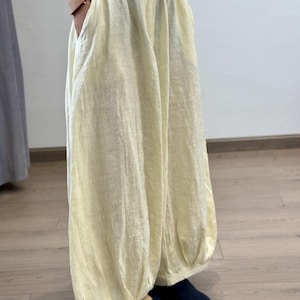 May include: Pale yellow linen pants with a gathered waist and pockets. The pants have a loose, flowing fit, with a gathered ankle. The fabric appears lightweight and breathable, suitable for warm weather. The pants are paired with dark blue slippers.