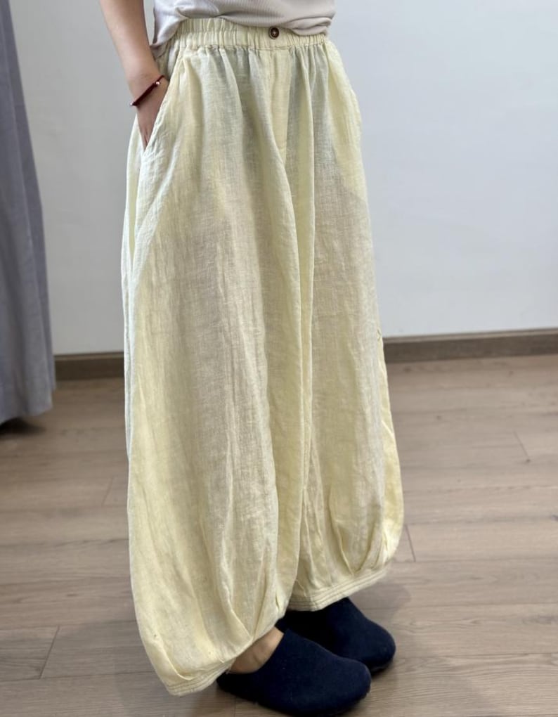 May include: Pale yellow linen pants with a gathered waist and pockets. The pants have a loose, flowing fit, with a gathered ankle. The fabric appears lightweight and breathable, suitable for warm weather. The pants are paired with dark blue slippers.
