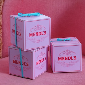 Puede incluir: Tres cajas cuadradas rosas con la palabra "Mendl's" en escritura roja. Cada caja está atada con una cinta azul claro. Las cajas están apiladas en una silla rosa.