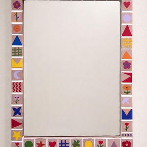 Peut inclure: Miroir rectangulaire avec une bordure décorative. La bordure est composée de petits carreaux carrés peints avec diverses formes et symboles colorés, notamment des fleurs, des étoiles et des motifs géométriques. Le miroir est fixé sur un mur blanc.