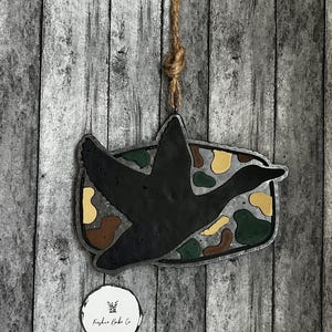 May include: A black duck-shaped ornament with a camouflage pattern and a jute string for hanging. The ornament is set against a weathered wooden background. A small white circular tag with text is at the bottom.