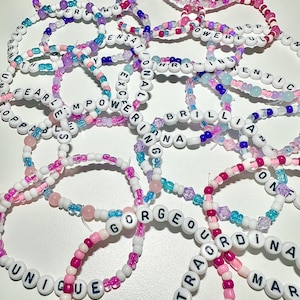 Pode incluir: Uma coleção de pulseiras de contas em várias cores, incluindo rosa, branco, roxo e azul. As pulseiras apresentam contas de letras brancas que soletram palavras como "BRAVE", "BEAUTIFUL" e "GORGEOUS".