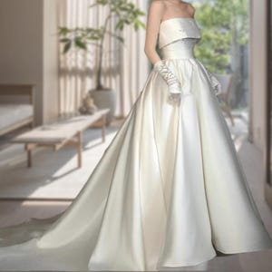 May include: A strapless ivory wedding gown with a full skirt and a long train. The bodice features delicate embroidery, and the ensemble includes matching gloves. The dress is made of a smooth, flowing fabric, creating an elegant silhouette.
