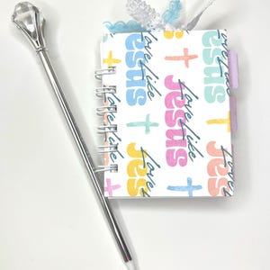 May include: A small spiral-bound notebook with a cover featuring the words "Love Like Jesus" and colorful crosses. A silver pen with a crystal-like top rests beside the notebook. The notebook has tabs and a decorative ribbon.