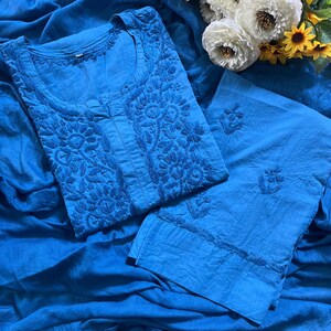 May include: A bright blue tunic and matching trousers set featuring intricate tonal floral embroidery on the bodice and sleeves. The fabric has a textured appearance, and the garment is displayed alongside a bouquet of white and yellow sunflowers.