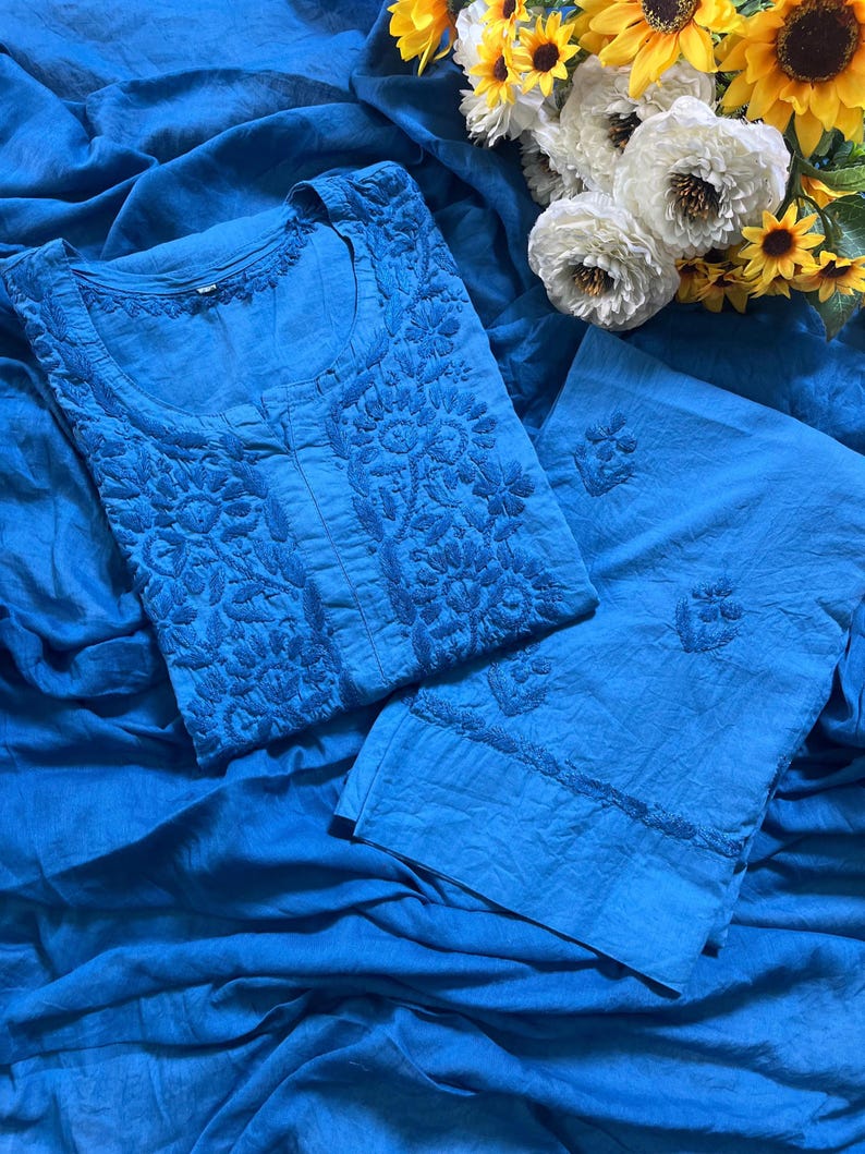 May include: A bright blue tunic and matching trousers set featuring intricate tonal floral embroidery on the bodice and sleeves. The fabric has a textured appearance, and the garment is displayed alongside a bouquet of white and yellow sunflowers.