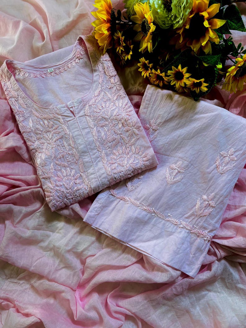 May include: A light pink cotton tunic and matching trousers set featuring intricate tonal floral embroidery. The tunic has a round neckline with a small centre slit, and the trousers feature embroidered floral motifs along the lower hem.