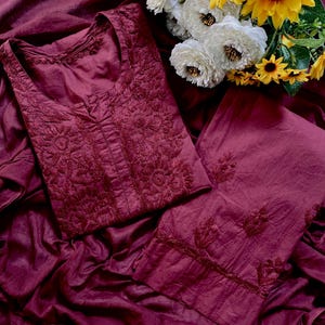 May include: A deep burgundy two-piece outfit consisting of a long-sleeved tunic and matching trousers. The tunic features intricate tonal embroidery around the neckline and chest, while the trousers have embroidered floral motifs at the hem.