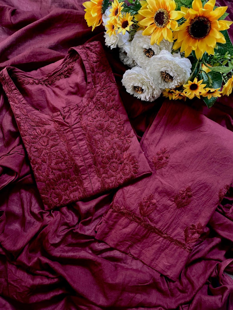 May include: A deep burgundy two-piece outfit consisting of a long-sleeved tunic and matching trousers. The tunic features intricate tonal embroidery around the neckline and chest, while the trousers have embroidered floral motifs at the hem.