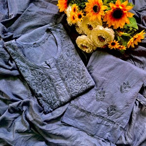 May include: A slate blue tunic and matching trousers set featuring intricate tonal embroidery on the bodice and scattered floral motifs on the pant legs, accompanied by a bouquet of bright yellow sunflowers and white carnations.