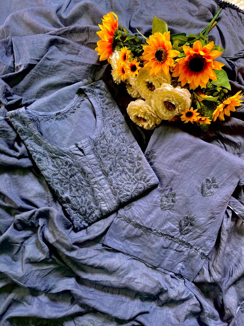 May include: A slate blue tunic and matching trousers set featuring intricate tonal embroidery on the bodice and scattered floral motifs on the pant legs, accompanied by a bouquet of bright yellow sunflowers and white carnations.