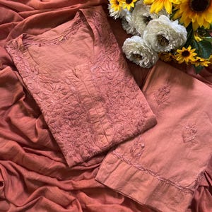 May include: A dusty rose-coloured tunic and matching trousers set featuring intricate tonal floral embroidery on the bodice and hem. The garment is crafted from a solid-coloured fabric and is displayed alongside a bouquet of yellow sunflowers and white flowers.