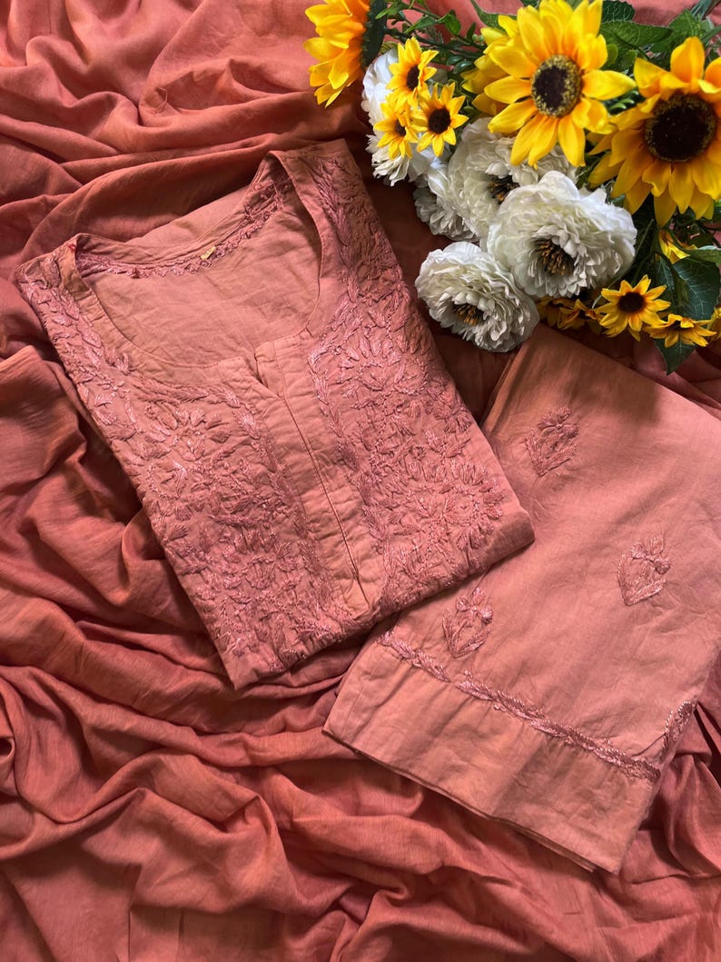 May include: A dusty rose-coloured tunic and matching trousers set featuring intricate tonal floral embroidery on the bodice and hem. The garment is crafted from a solid-coloured fabric and is displayed alongside a bouquet of yellow sunflowers and white flowers.