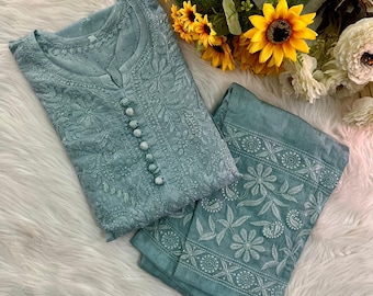 Puede incluir: Un conjunto de kurta y pantalón bordado de color verde azulado claro. La kurta presenta una botonadura frontal y un intrincado bordado floral. Los pantalones también tienen patrones florales bordados. Un ramo de girasoles y flores blancas descansa junto a la ropa.