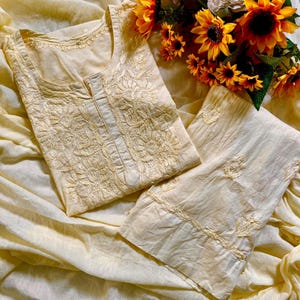 May include: A pale yellow tunic and matching trousers set featuring intricate tonal embroidery on the bodice and cuffs. The garment has a round neckline with a small centre slit and is displayed alongside a bouquet of bright yellow sunflowers.