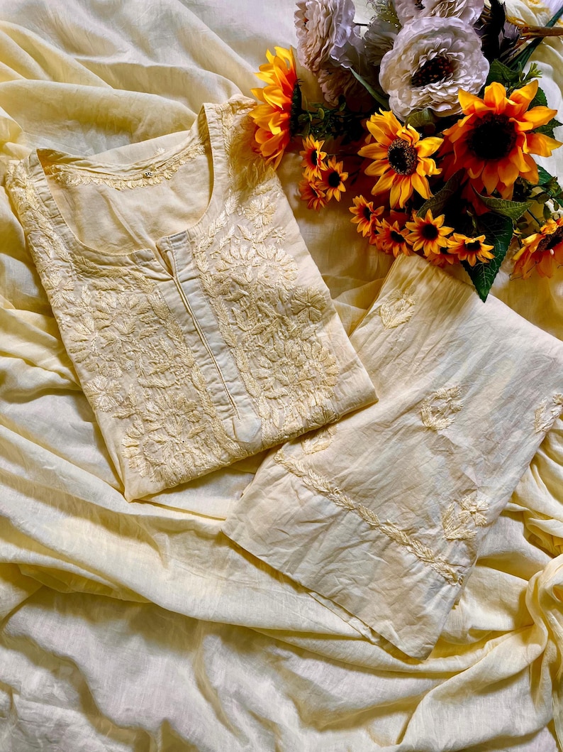 May include: A pale yellow tunic and matching trousers set featuring intricate tonal embroidery on the bodice and cuffs. The garment has a round neckline with a small centre slit and is displayed alongside a bouquet of bright yellow sunflowers.