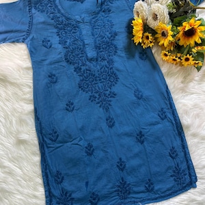 May include: A blue tunic featuring intricate tonal embroidery around the neckline and scattered floral motifs across the front. The garment has long sleeves and a round neckline, complemented by a decorative bouquet of white and yellow flowers.