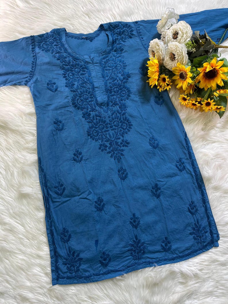 May include: A blue tunic featuring intricate tonal embroidery around the neckline and scattered floral motifs across the front. The garment has long sleeves and a round neckline, complemented by a decorative bouquet of white and yellow flowers.