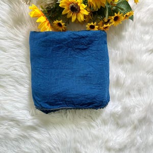 May include: A folded square of deep blue fabric with a visible textured weave and a subtle dark trim along the bottom edge. The textile is positioned below a cluster of bright yellow sunflowers and white flowers with green leaves and stems.