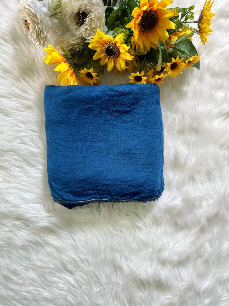 May include: A folded square of deep blue fabric with a visible textured weave and a subtle dark trim along the bottom edge. The textile is positioned below a cluster of bright yellow sunflowers and white flowers with green leaves and stems.