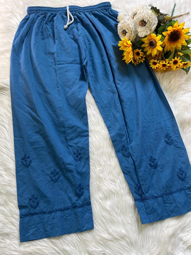 May include: Blue wide-leg trousers featuring an elastic waistband with a white drawstring and tonal floral embroidery scattered across the lower legs, finished with a horizontal stitched hem detail near the bottom of each trouser leg.